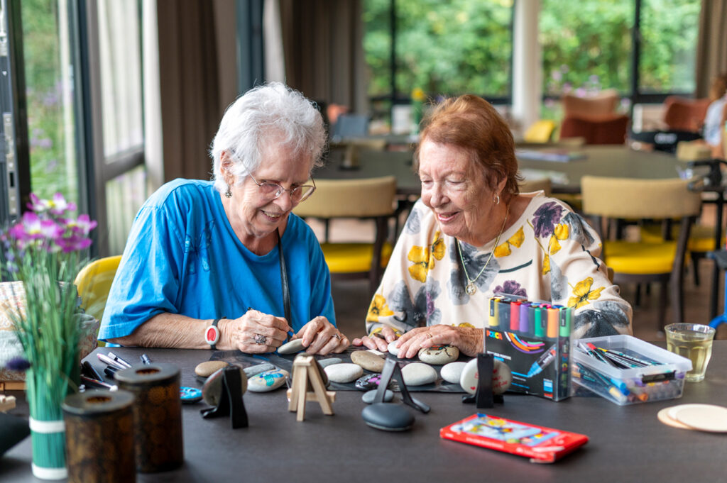 Twee cliënten zijn bezig met het beschilderen van stenen aan een lange tafel in de ontmoetingsruimte